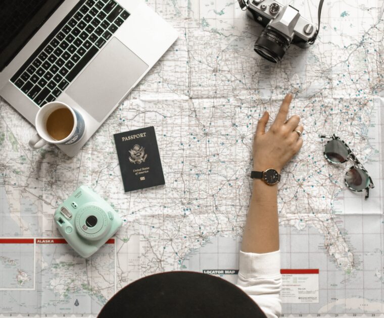 Traveler pointing to a map of the United States. With a passport, camera, sunglasses, and laptop scattered on the map.